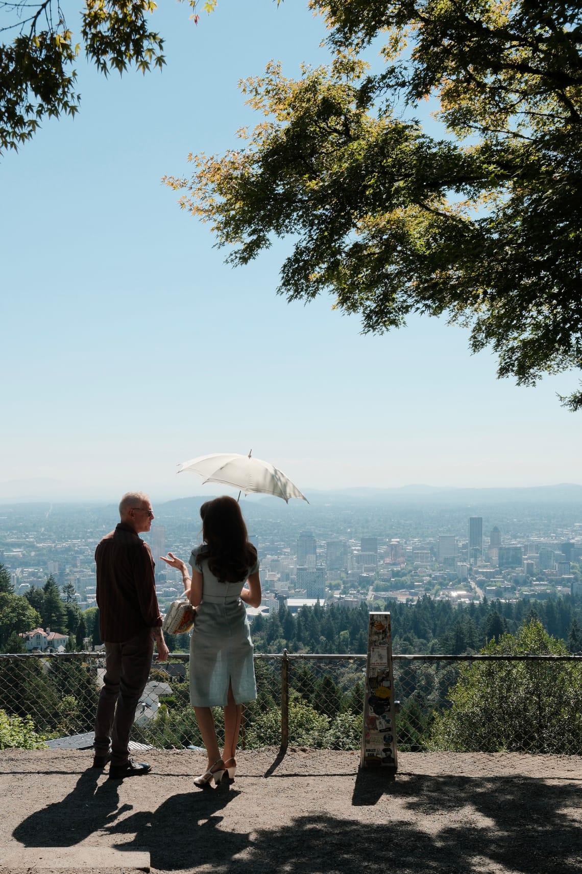 A woman with a parasol talks to a man with the Portland skyline before them. 