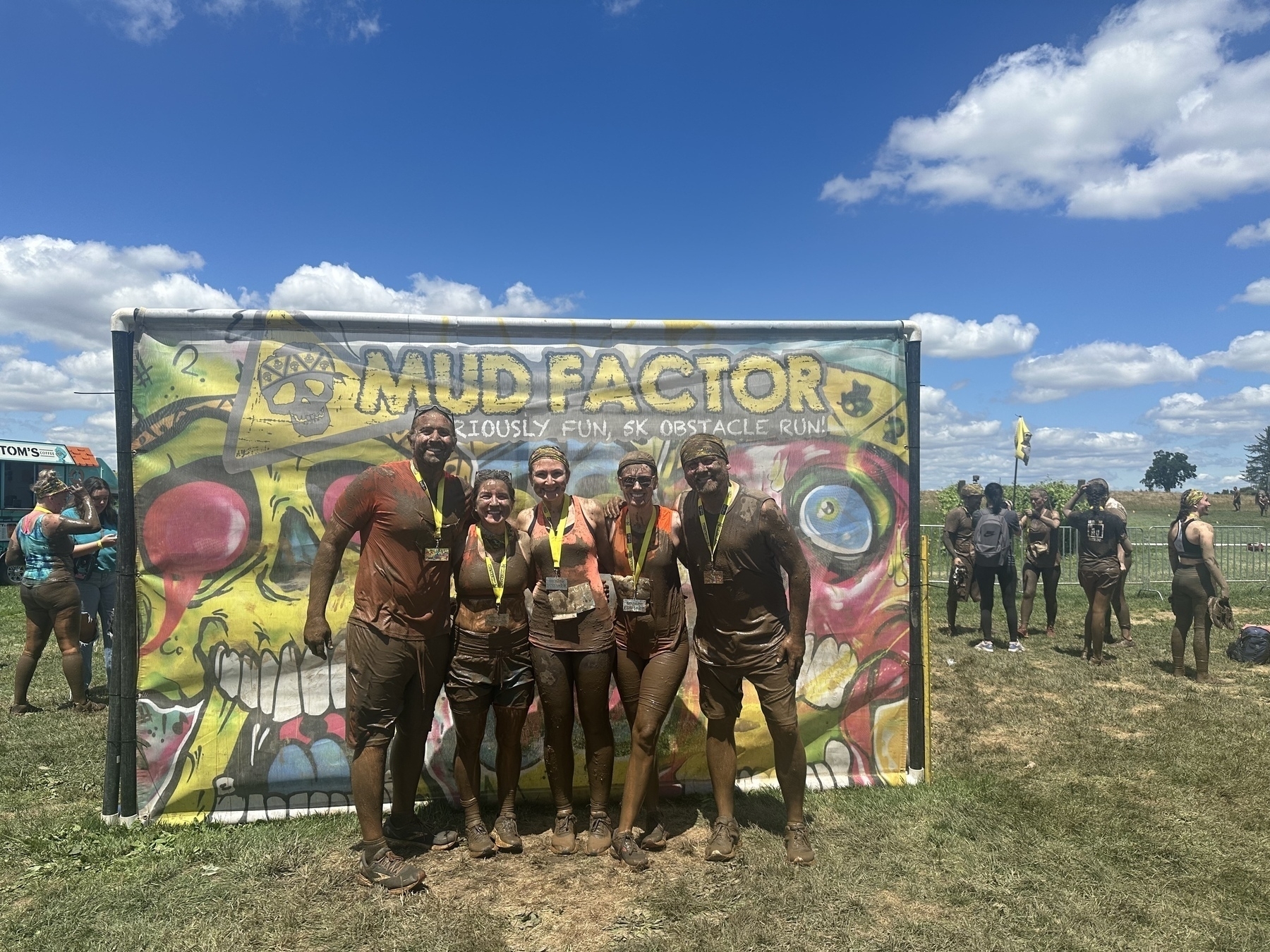 Five people covered in mud pose in front of a colorful backdrop for "Mud Factor," an obstacle run event.