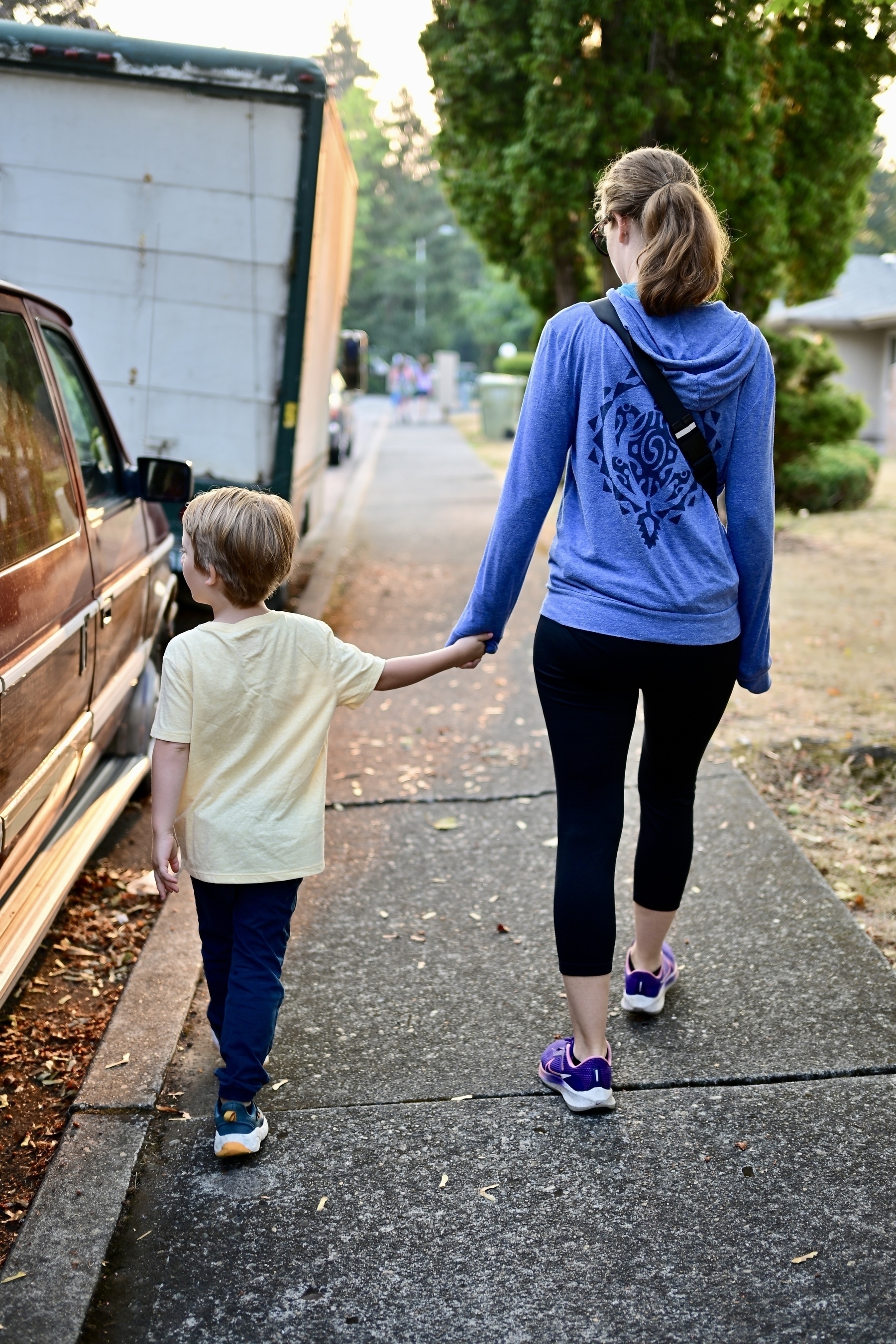 A woman wearing a blue hoodie and black leggings walks hand in hand with a child on a sidewalk.