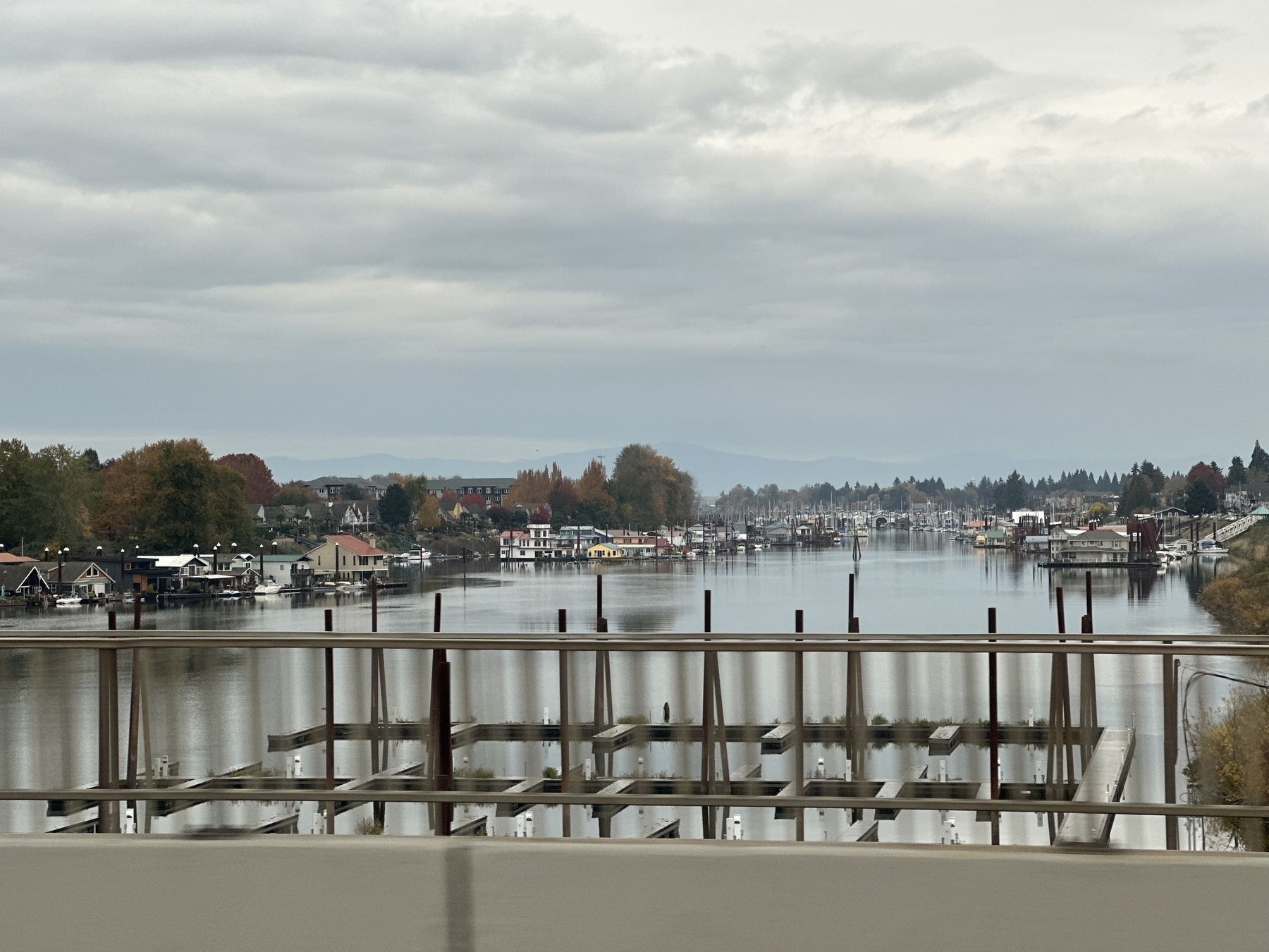 A calm river is lined with houses and trees under an overcast sky, with dock structures visible in the foreground.