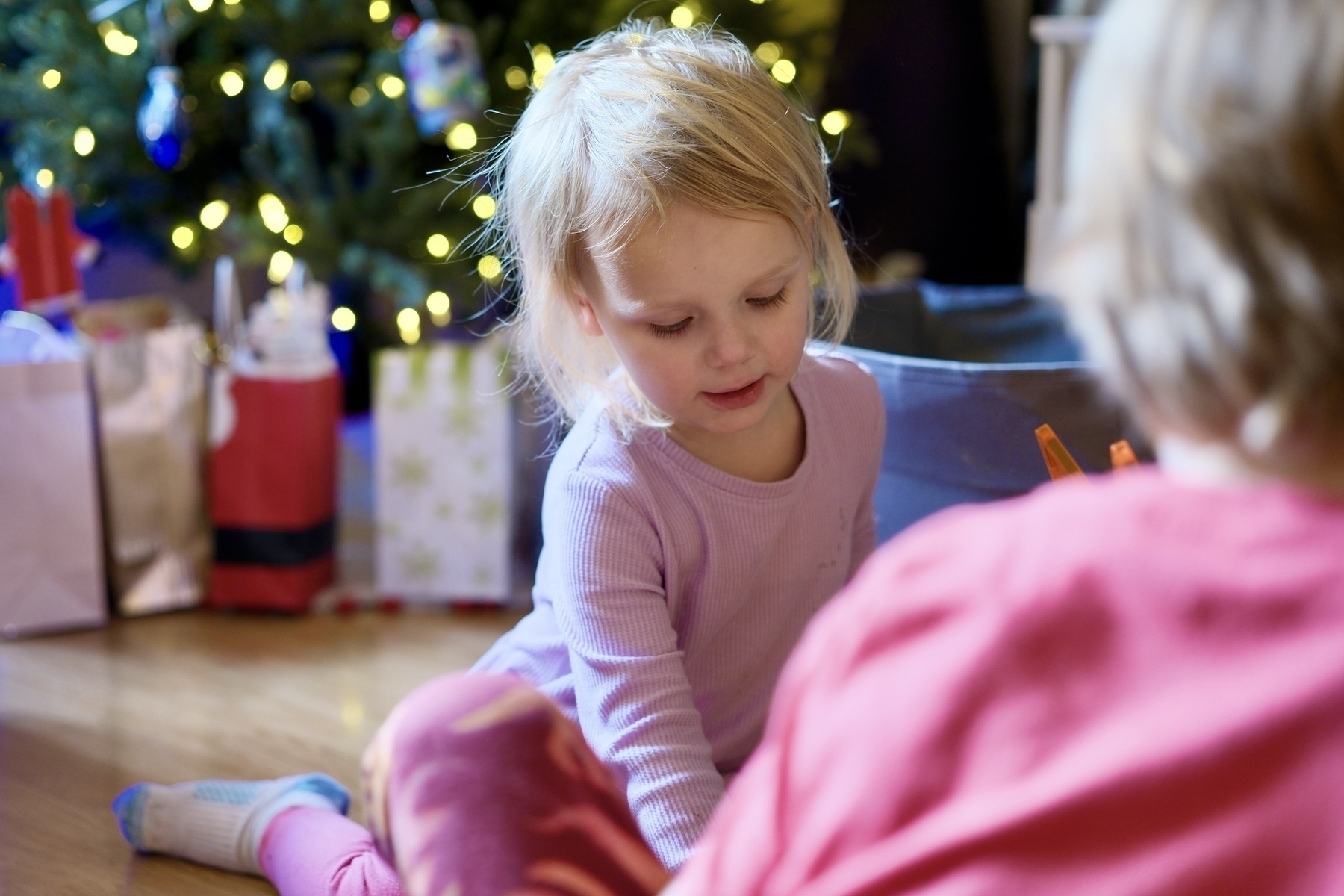 A young child plays on the floor near a decorated Christmas tree with presents underneath.