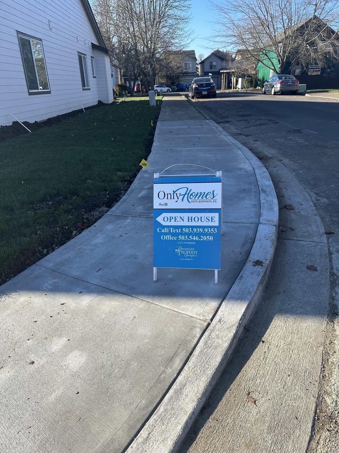 A real estate sign for an open house is placed on a sidewalk in a suburban neighborhood with an agency name of “OnlyHomes”. 