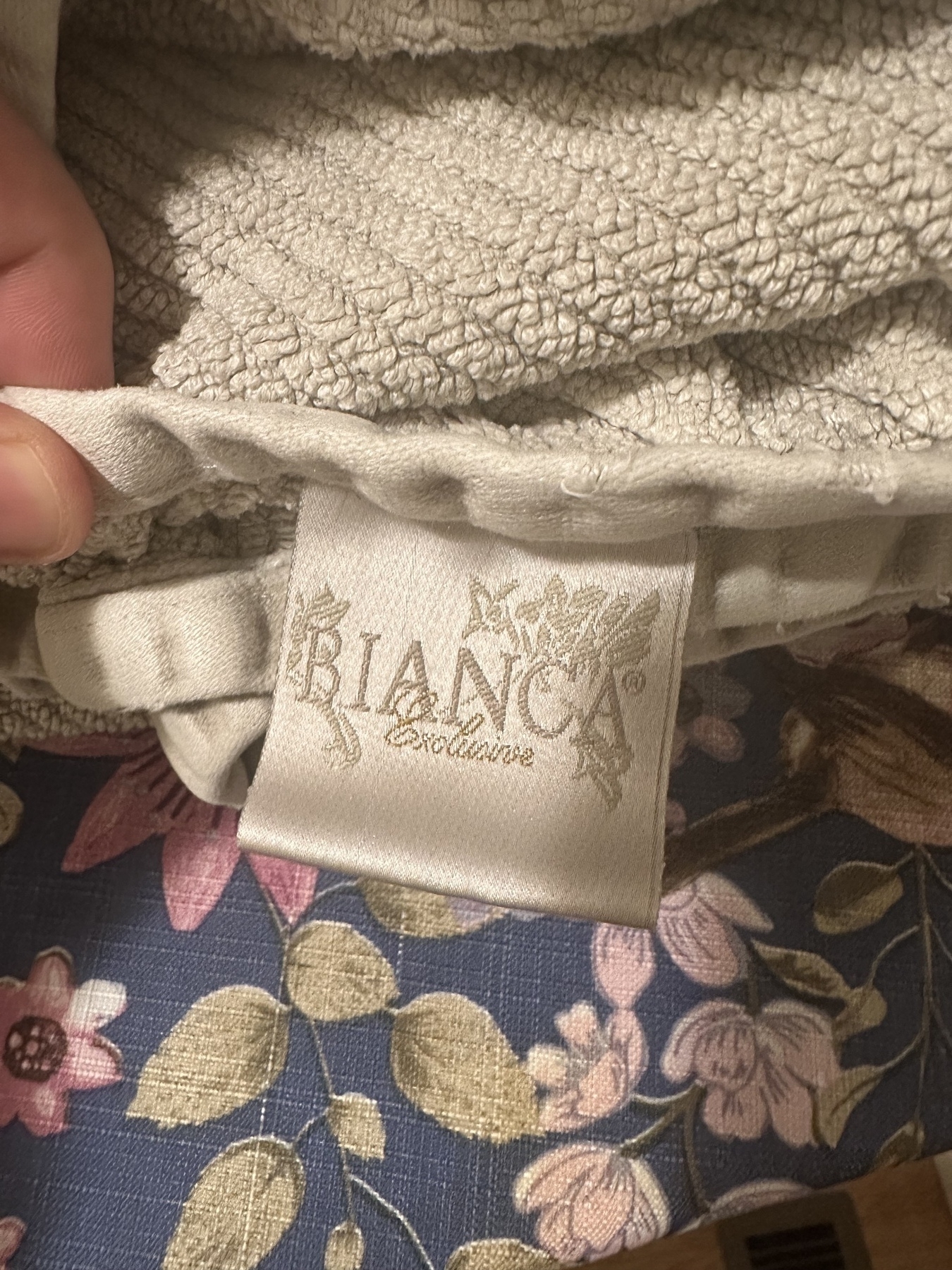 A close-up of a beige, textured towel with a "BIANCA Exclusive" label showing above a floral-patterned surface.