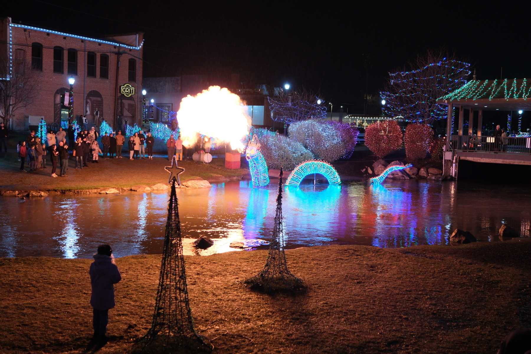 Auto-generated description: A nighttime scene shows festive lights, people gathered by a pond, and a fire performer creating a dramatic burst of flame.